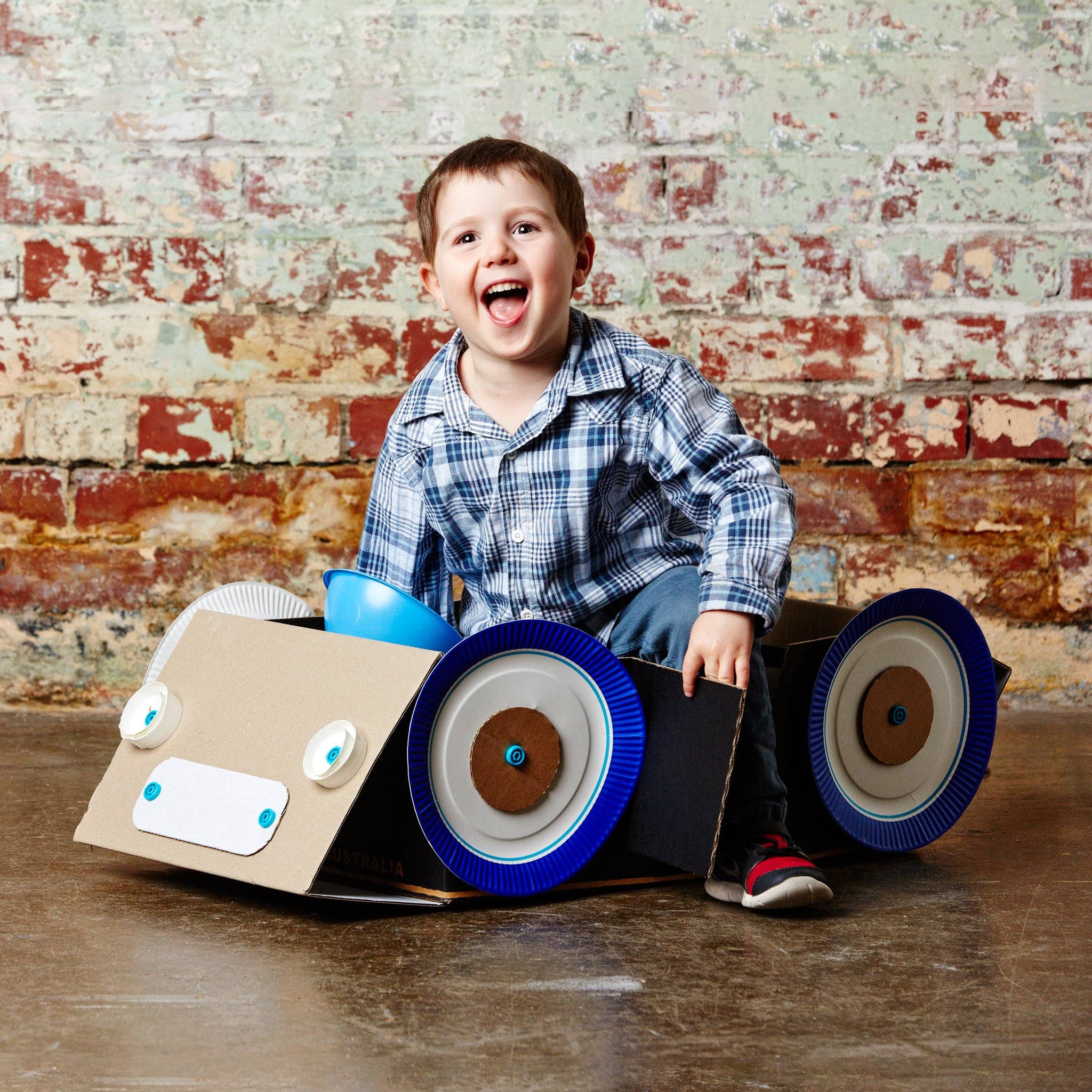 Child in a cardboard car with blue paper plate wheels and bottle cap lights, held together by small blue screws.