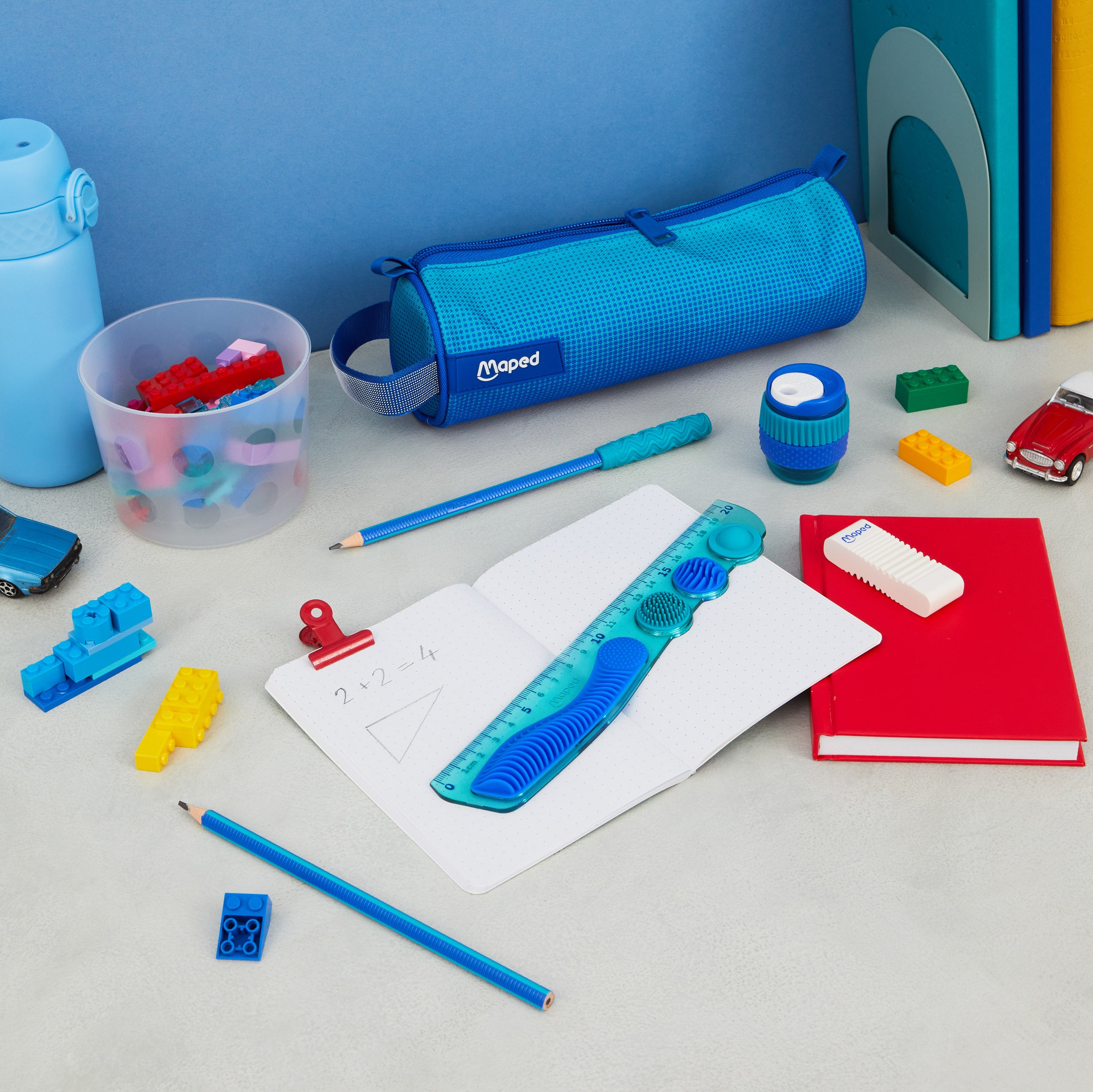 Blue stationery set featuring a textured sensory ruler, pencil case, and pencils on a desk with colorful toys.