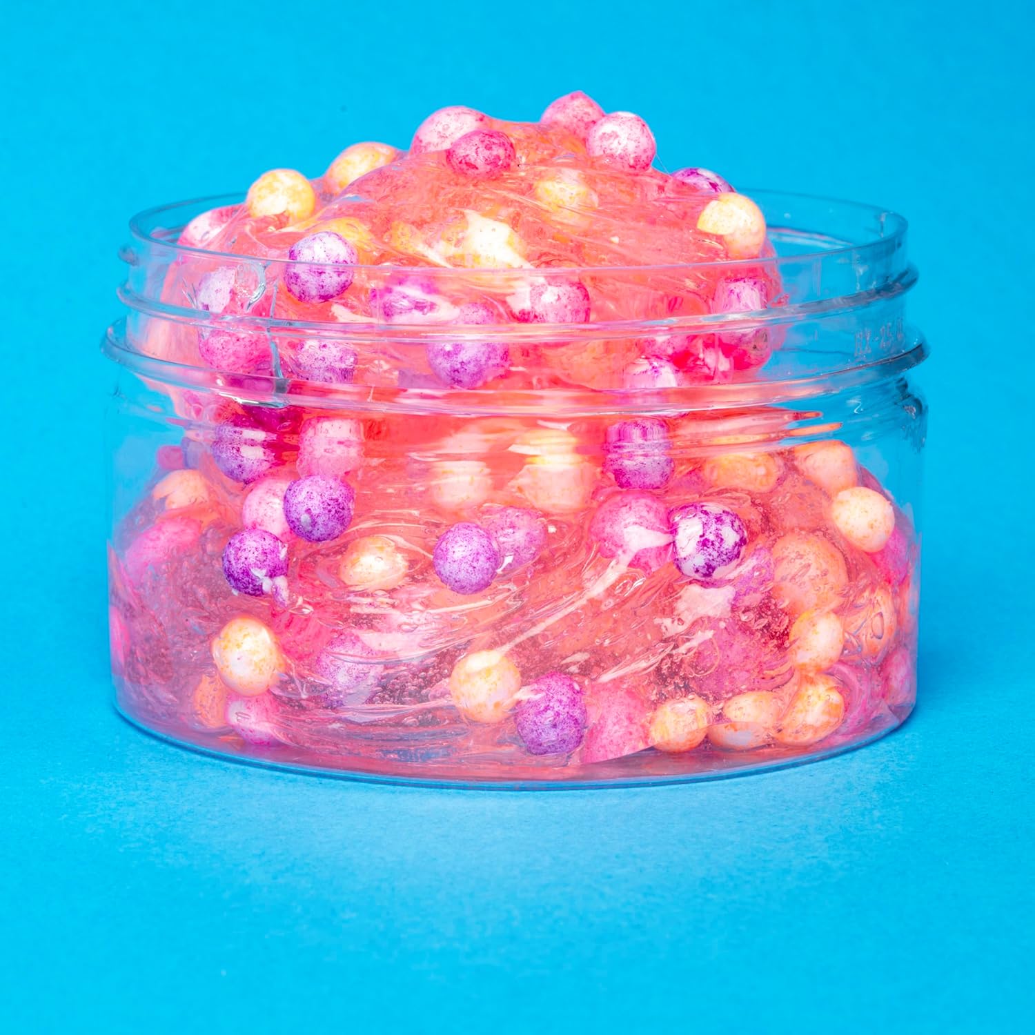 Pink translucent slime with yellow, pink, and purple foam beads in a clear jar against a bright blue background.