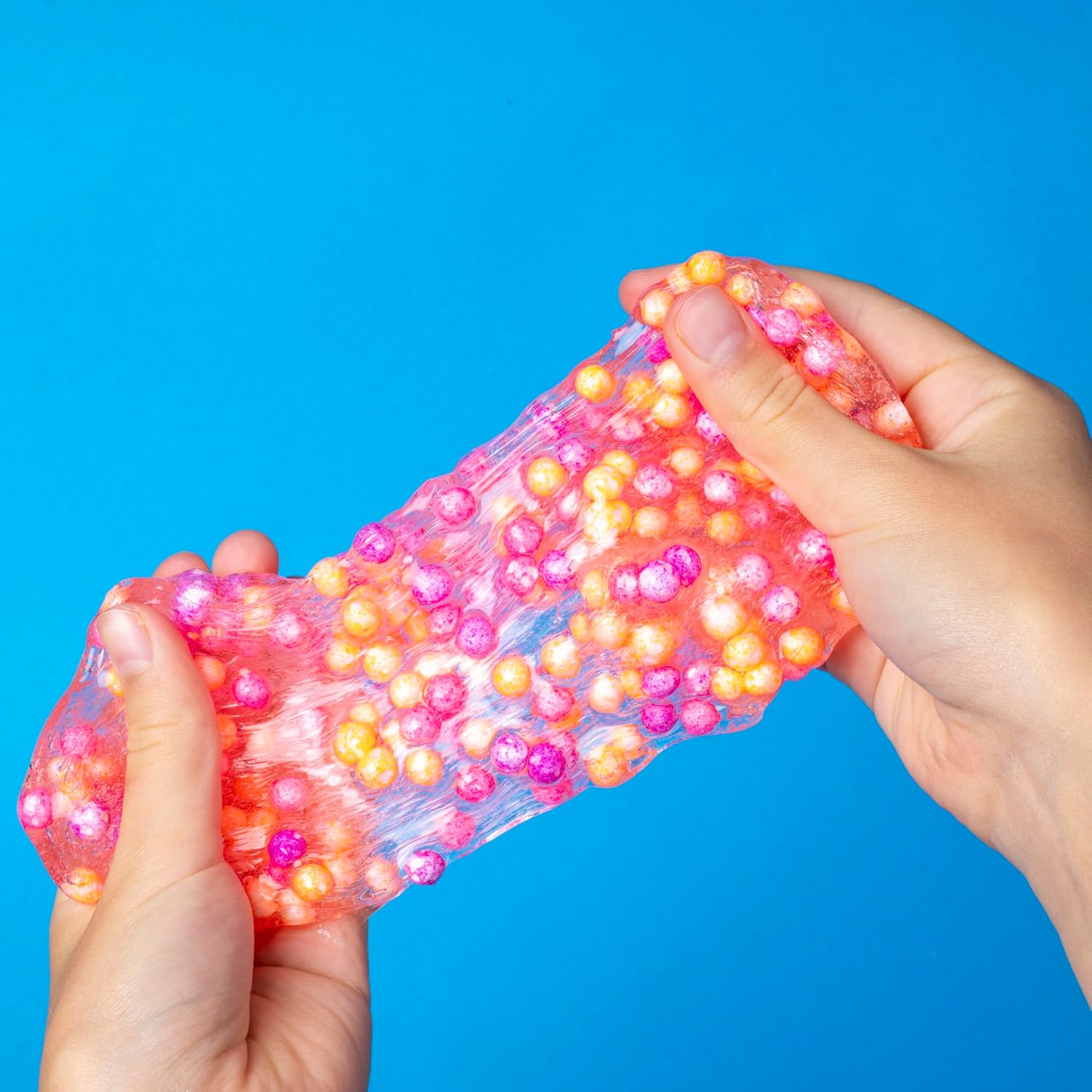 Two hands stretch pink translucent slime filled with colorful crunchy foam beads against a bright blue background.