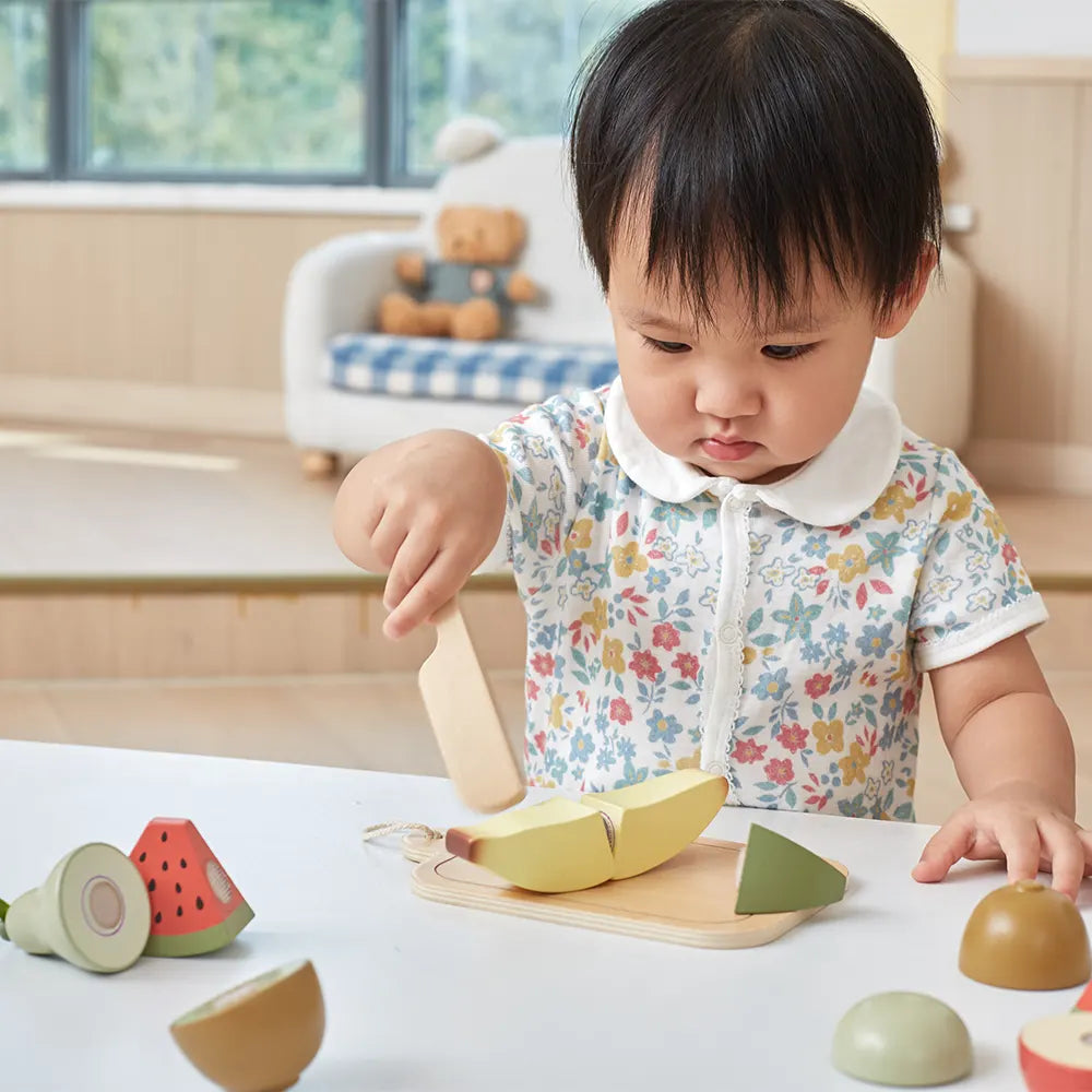 Toddler slices a yellow wooden banana with a toy knife on a board; colorful fruit pieces sit on a white table.