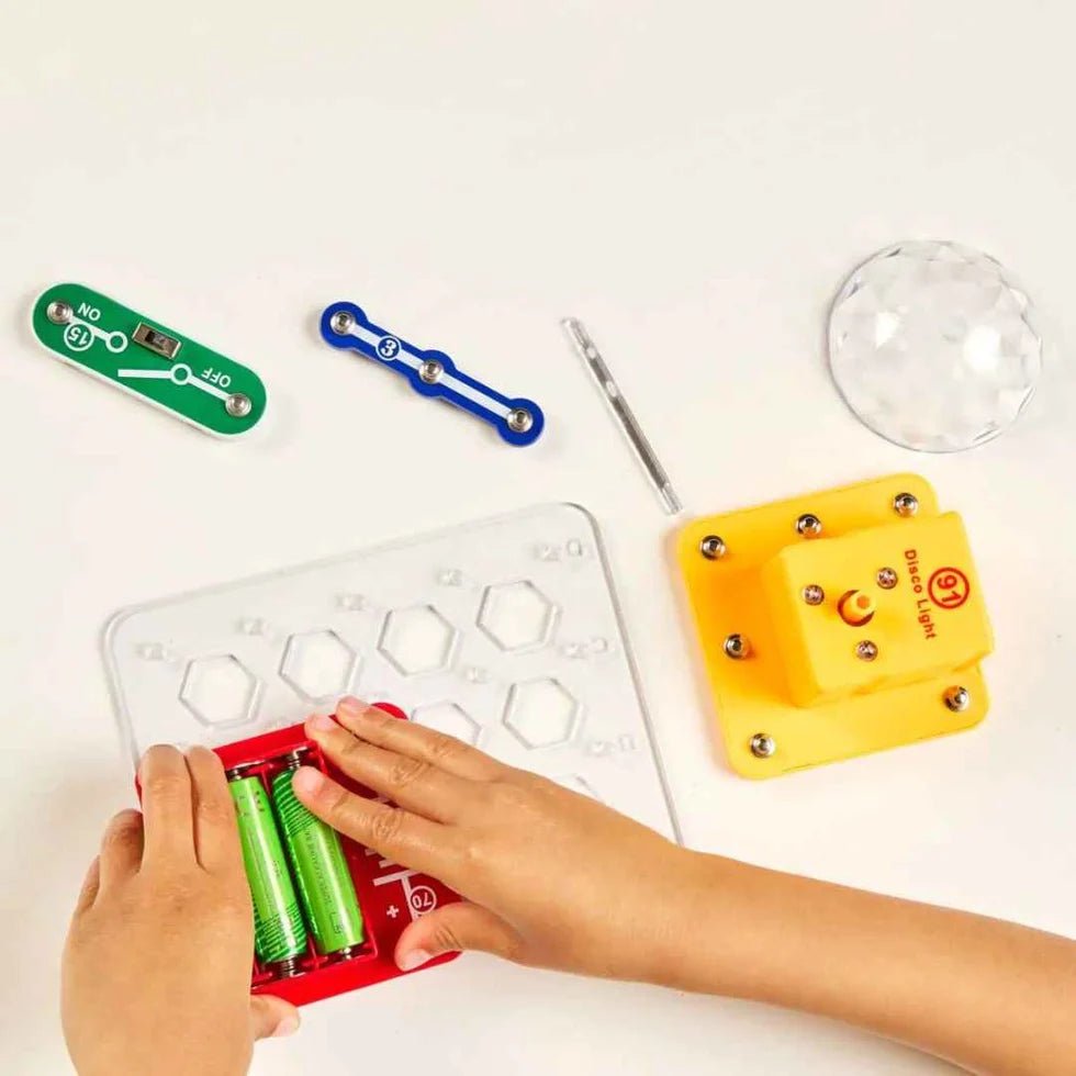 Child assembling an electronic kit with a red battery pack, yellow disco light motor, green switch, and clear dome.