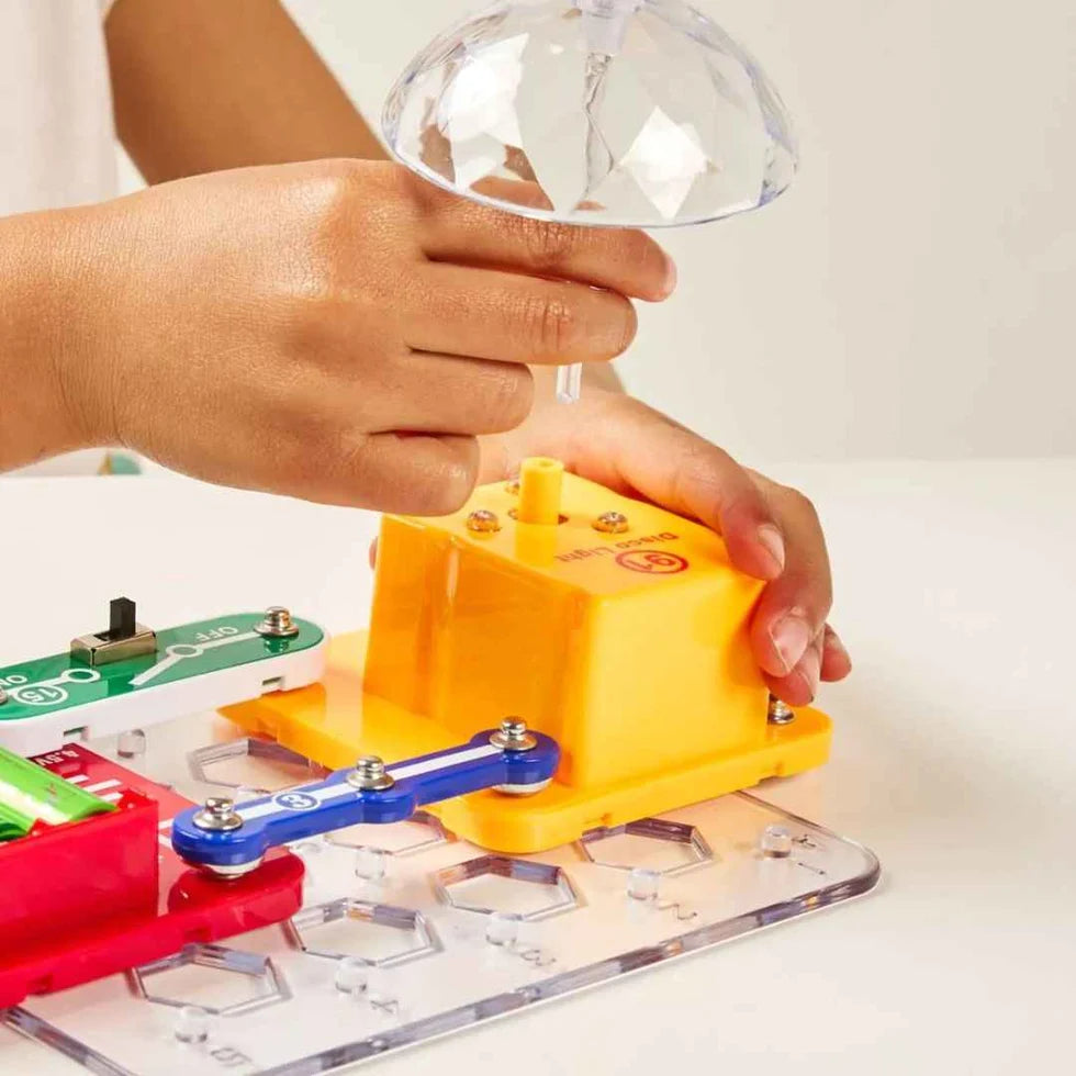 Child's hands attaching a clear disco light dome to a yellow base on a colorful electronic snap circuit toy.