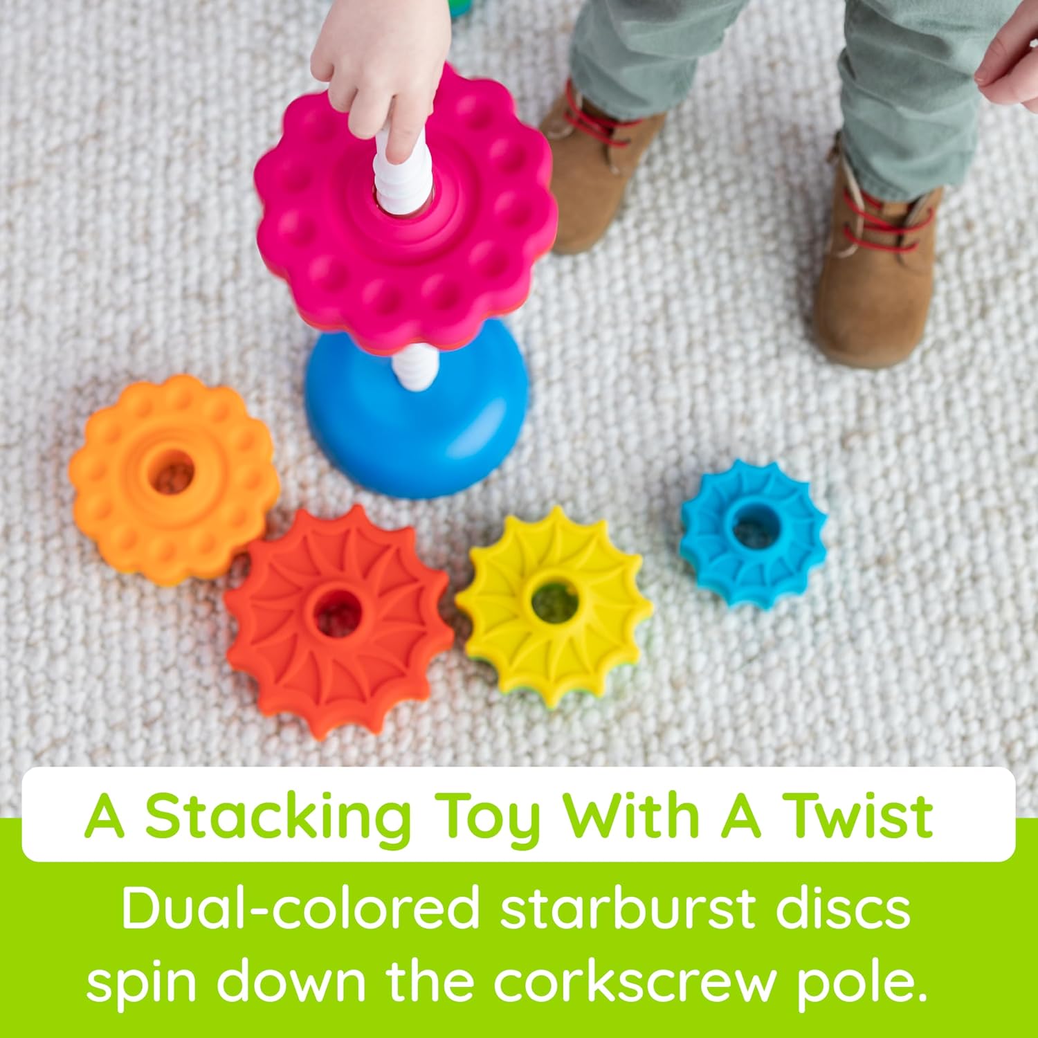 A child's hand stacks a pink starburst disc on a white spiral pole. Colorful discs are scattered on a carpet.