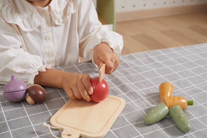 Child cuts a red wooden tomato on a board next to purple, brown, and green toy vegetables on a grid tablecloth.