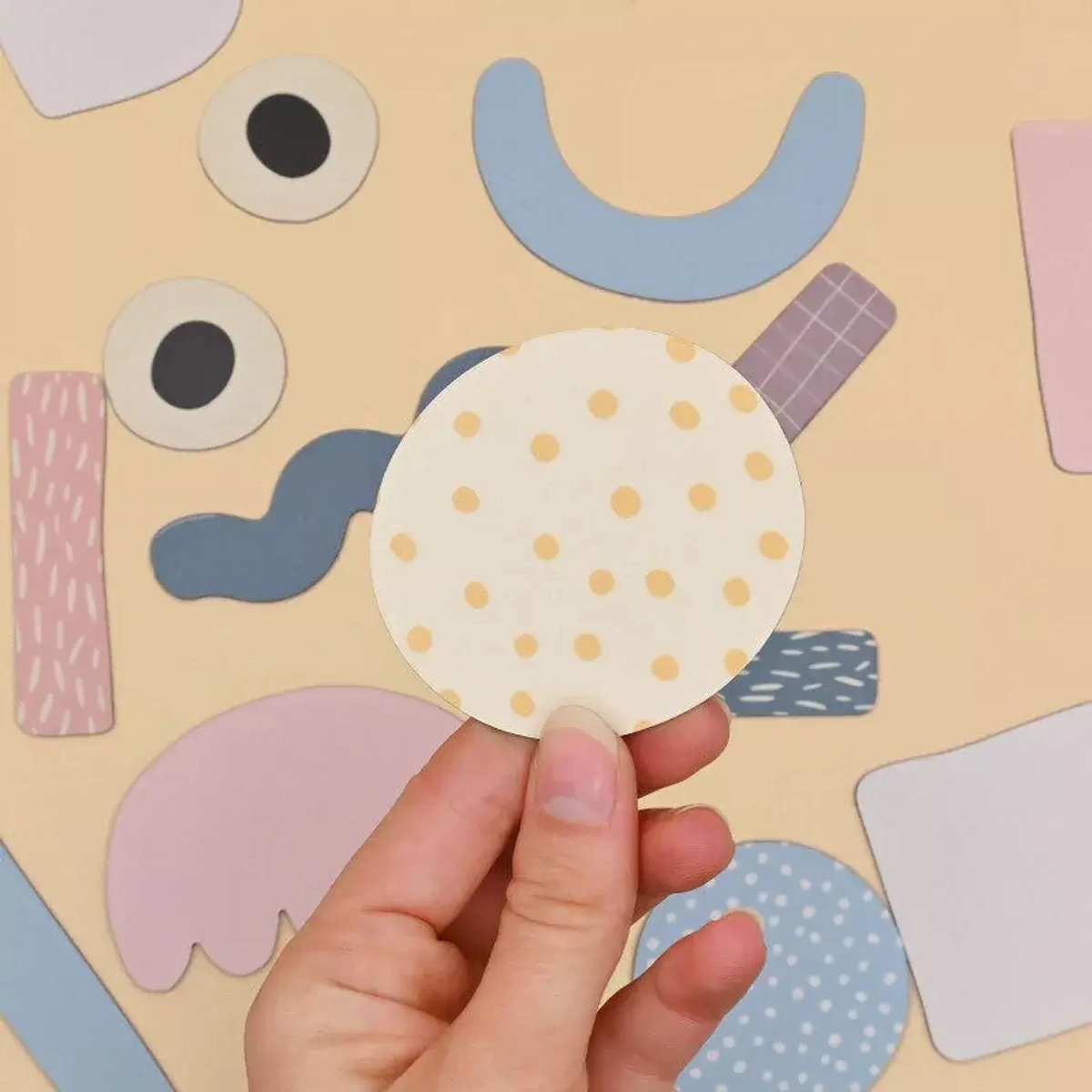 Hand holds a cream circular magnet with yellow dots over colorful, abstract-shaped magnets on a beige background.