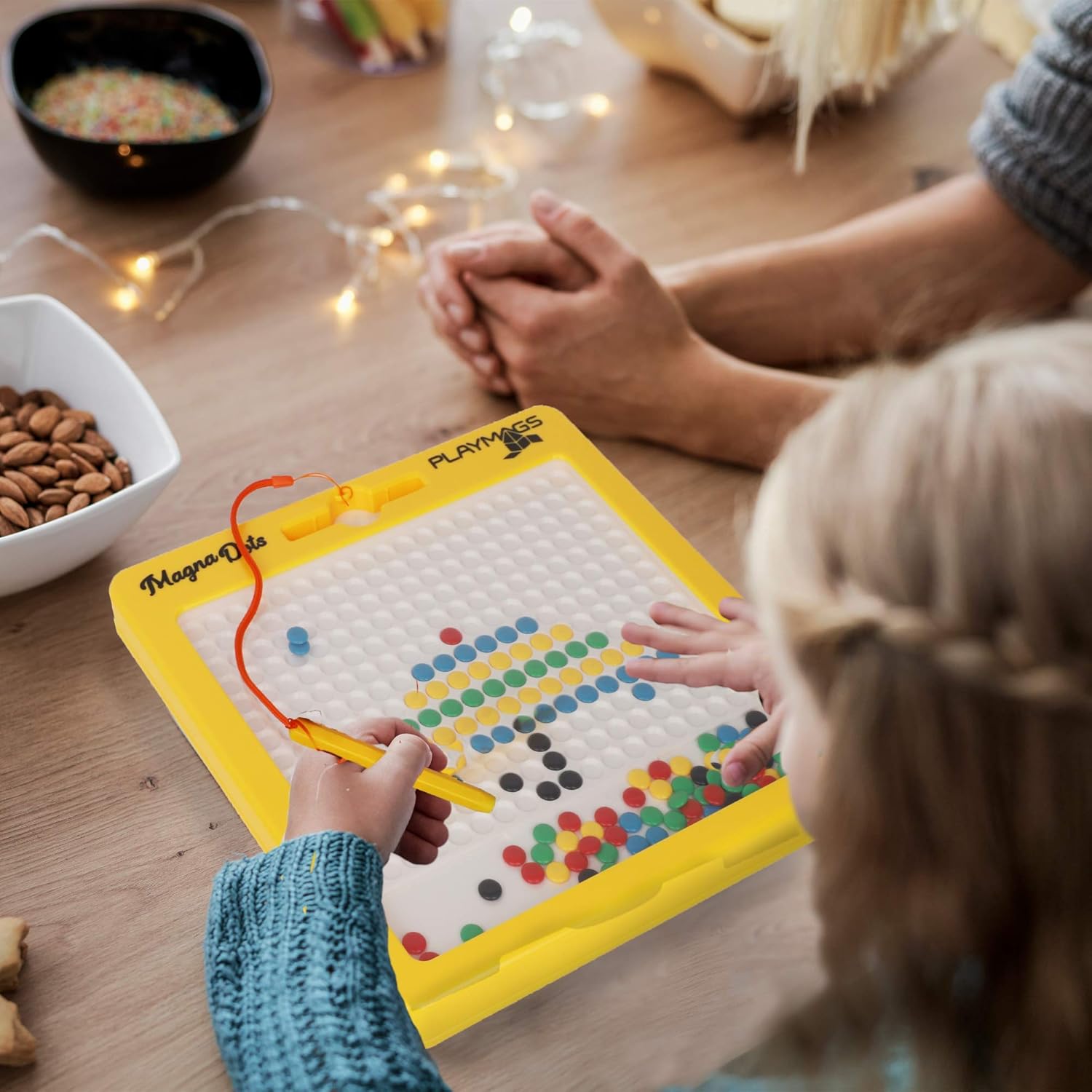 Child using an orange magnetic pen to move colorful dots on a yellow drawing board to create a mosaic pattern.