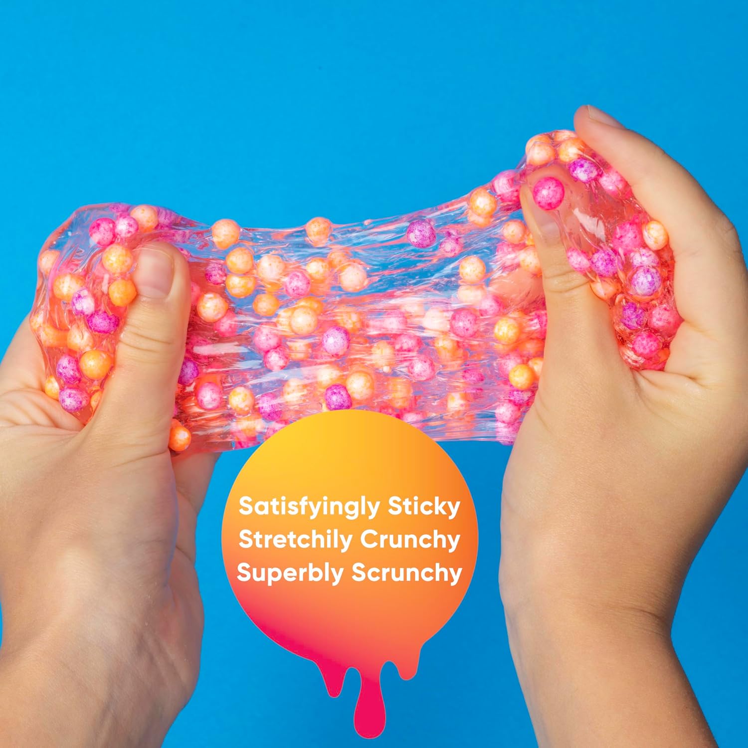 Hands stretching clear slime filled with pink and orange foam beads against a bright blue background.