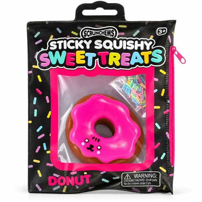 A pink iced squishy donut toy with a cute face inside black packaging with a small bag of colorful sprinkles.