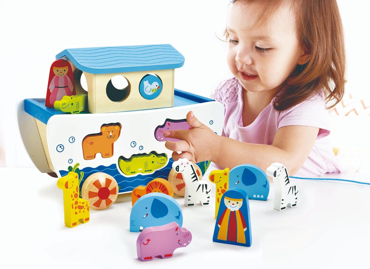 Toddler playing with a colorful wooden boat shape sorter and matching animal figurines on a white background.