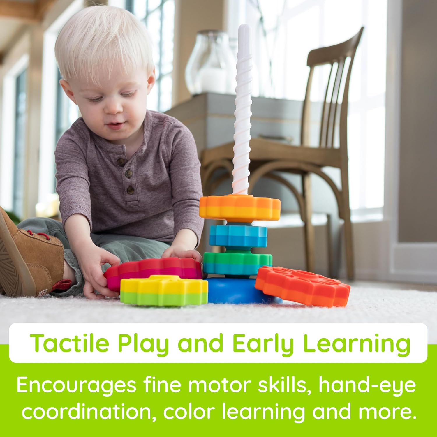 A toddler boy sits on a rug stacking bright, multi-colored gear-shaped discs onto a tall white corkscrew pole.