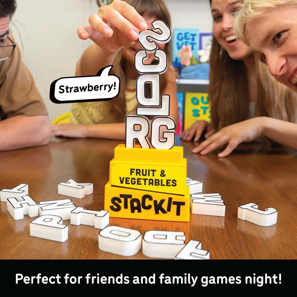 A group of people gathered around a wooden table playing with the Stackit Fruit & Vegetables word game. The bright yellow game base is stacked with white letter tiles, and one person is holding a tile.