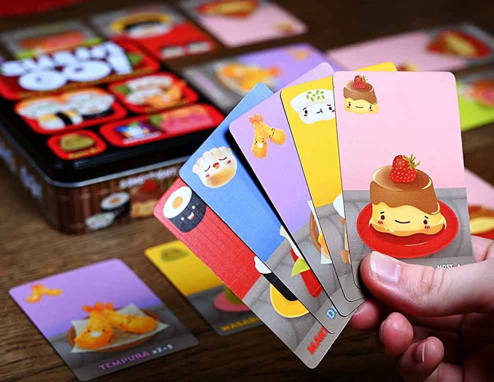 A hand holds a fan of colorful illustrated cards featuring cute food illustrations, with a matching game tin and additional cards blurred in the background on a wooden surface.